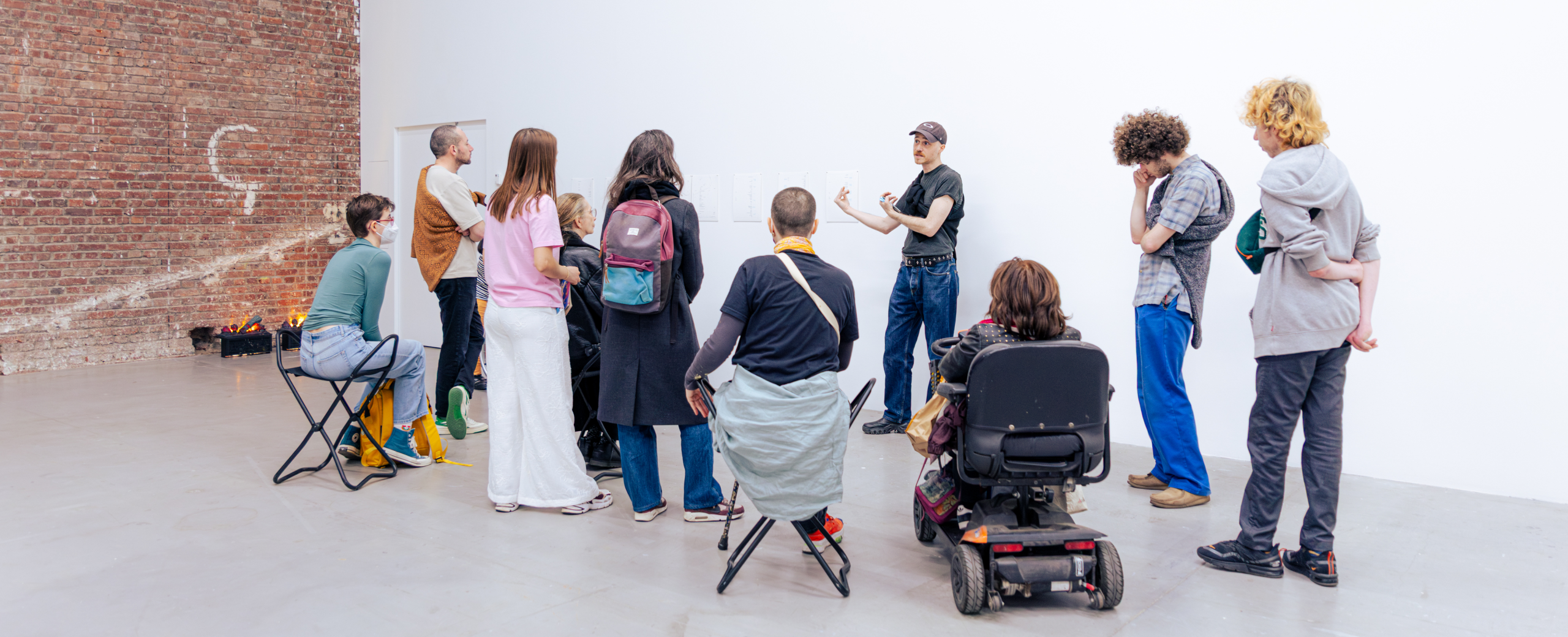 Un groupe de personnes écoute un intervenant dans une galerie. Certaines sont assises, d'autres debout, et l'une d'entre elles se déplace en fauteuil roulant. Le lieu est lumineux, avec des murs en briques et des murs blancs.