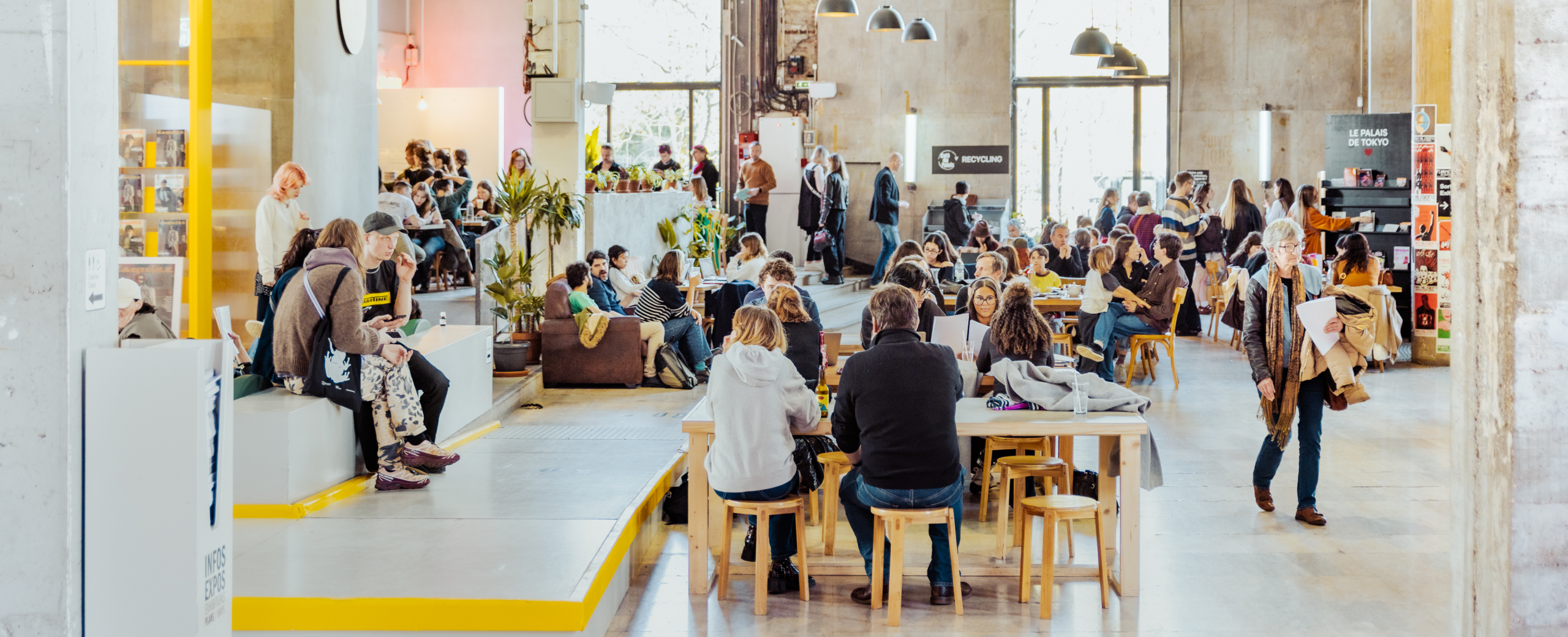 Un grand café spacieux où de nombreuses personnes sont assises aux tables, discutent ou travaillent ; la lumière naturelle pénètre à flots par les hautes fenêtres, et des plantes agrémentent l'espace.