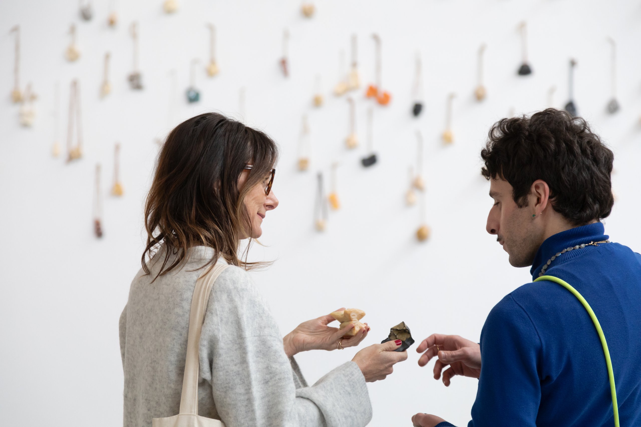 Deux personnes se tiennent devant un mur blanc sur lequel sont accrochés de petits objets. Elles discutent, l'une d'elles tenant quelques objets à la main.