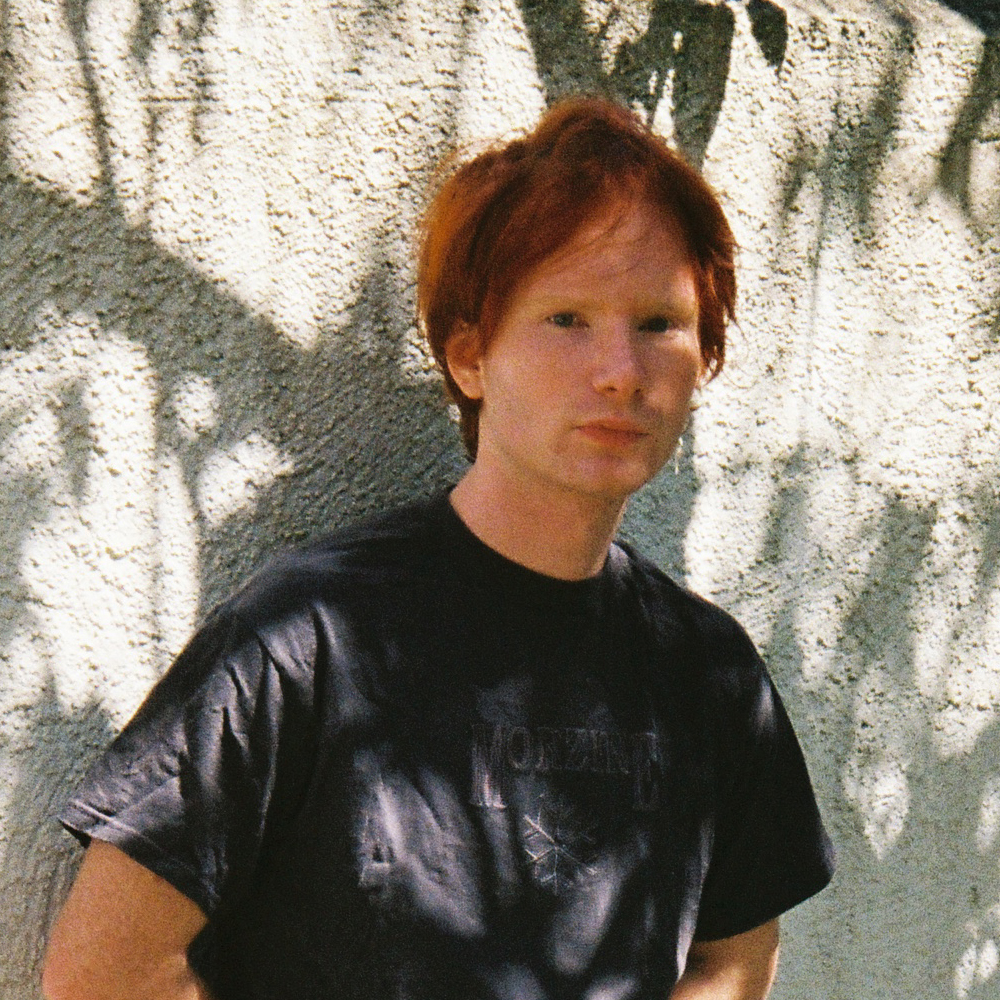 Une personne aux cheveux roux, vêtue d'un t-shirt noir, se tient devant un mur texturé, baigné par la lumière du soleil et les ombres.