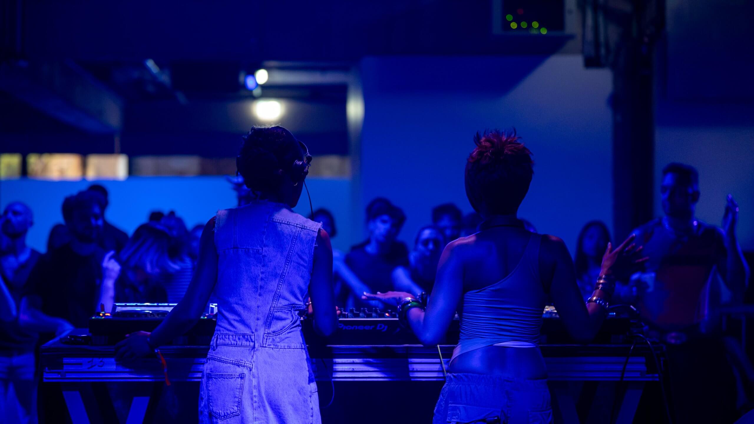 Deux DJ aux platines dans une boîte de nuit faiblement éclairée, aux tons bleutés.