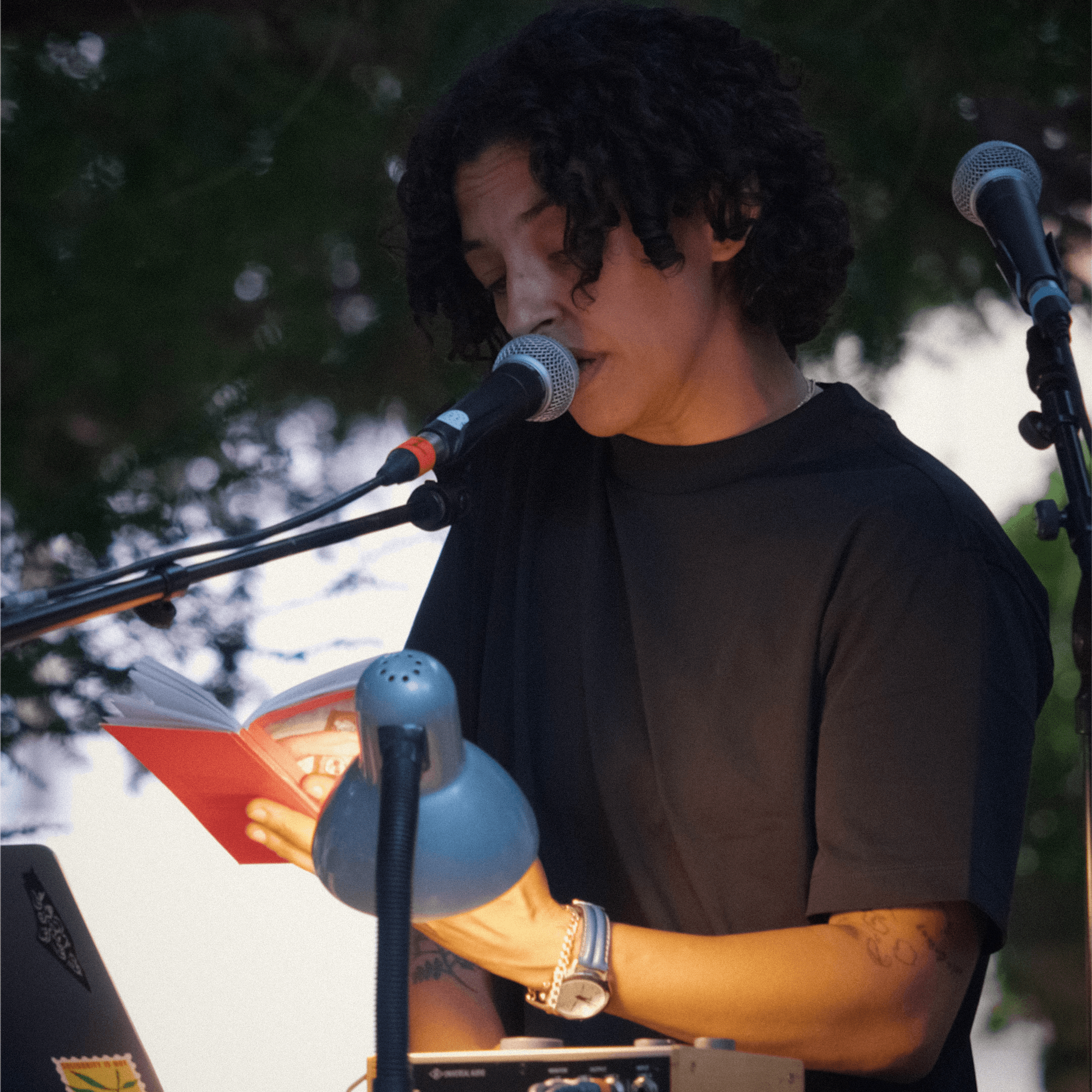 A person with curly hair reading from a small book into a microphone, illuminated by a desk lamp.