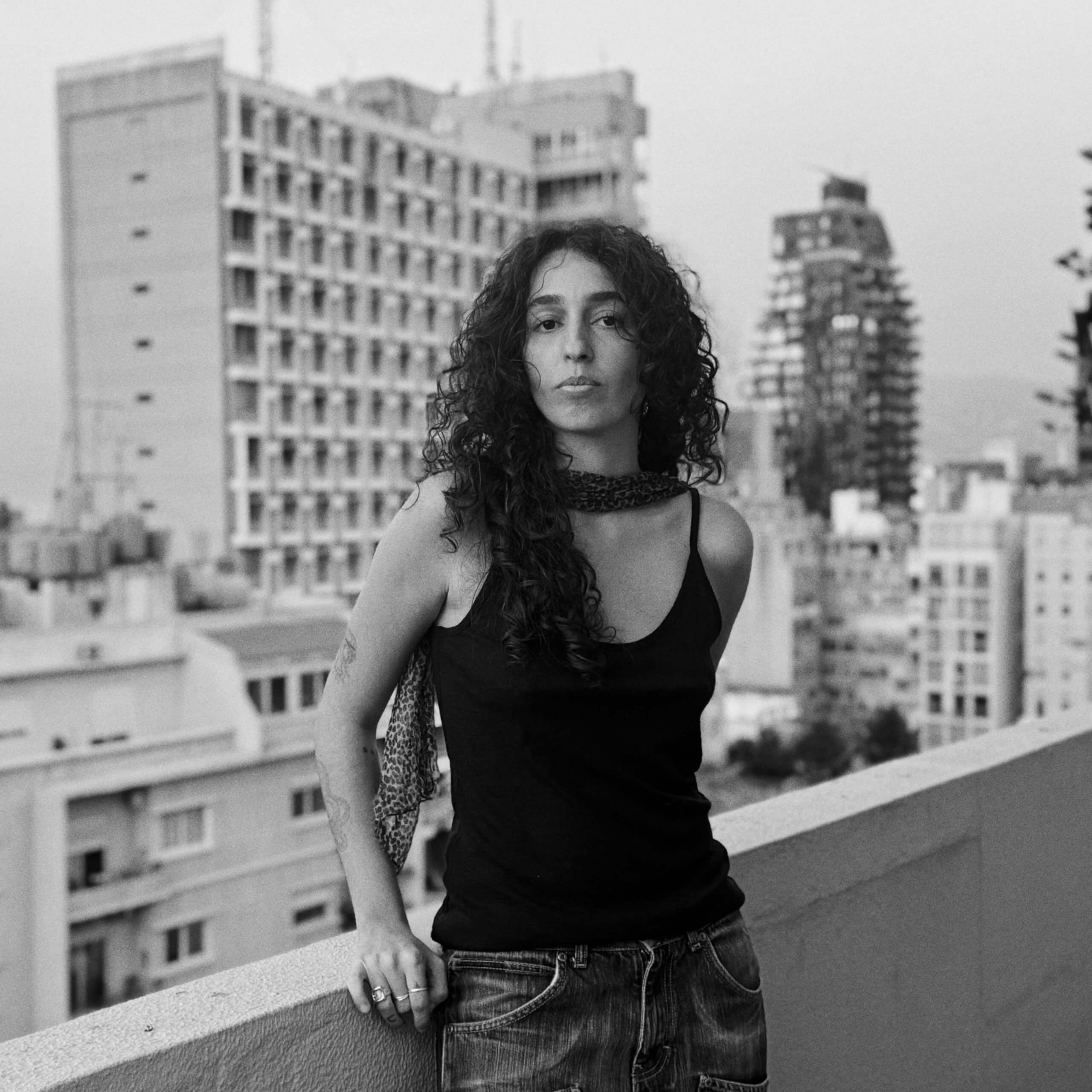 A woman with long curly hair wearing a tank top and jeans stands on a balcony overlooking a city skyline.